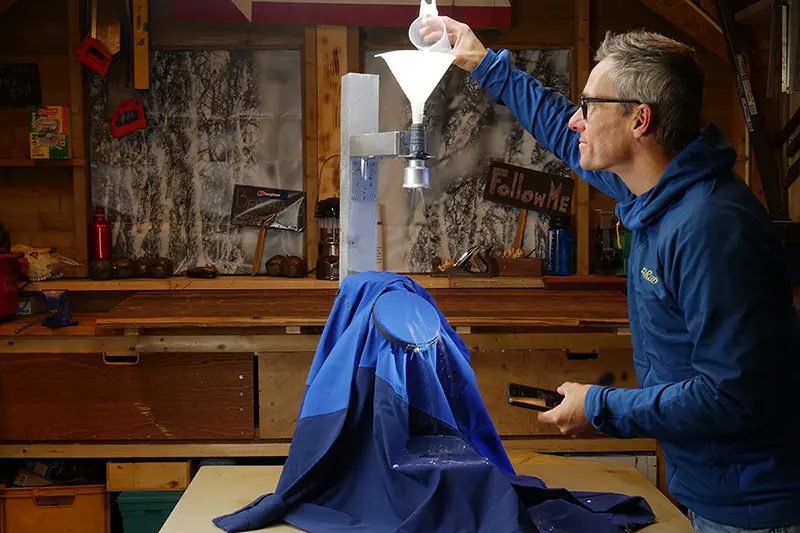 In een werkplaats giet een man via een trechter water op een blauwe waterdichte jas, waarmee hij de waterafstotende eigenschappen ervan demonstreert: het water vormt parels en loopt van de stof af.